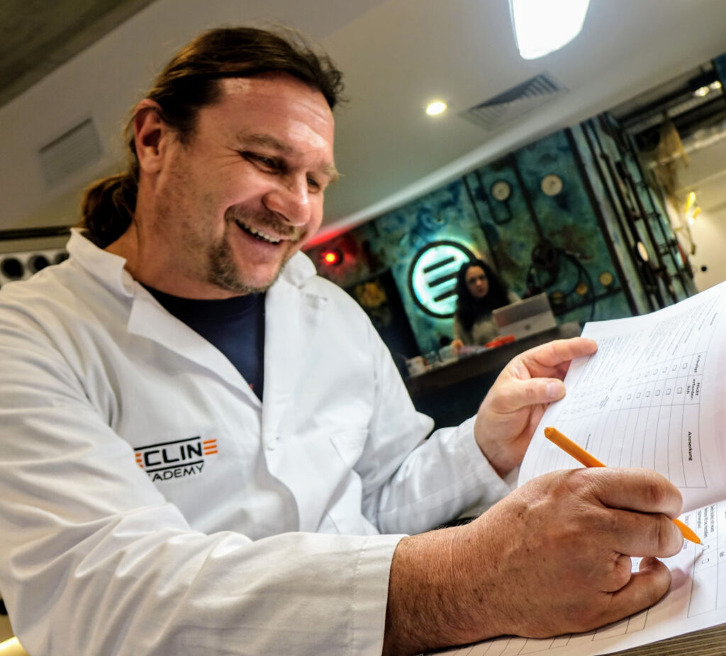 Mann in weißem Kittel lächelt und schreibt mit Bleistift in ein Formular.